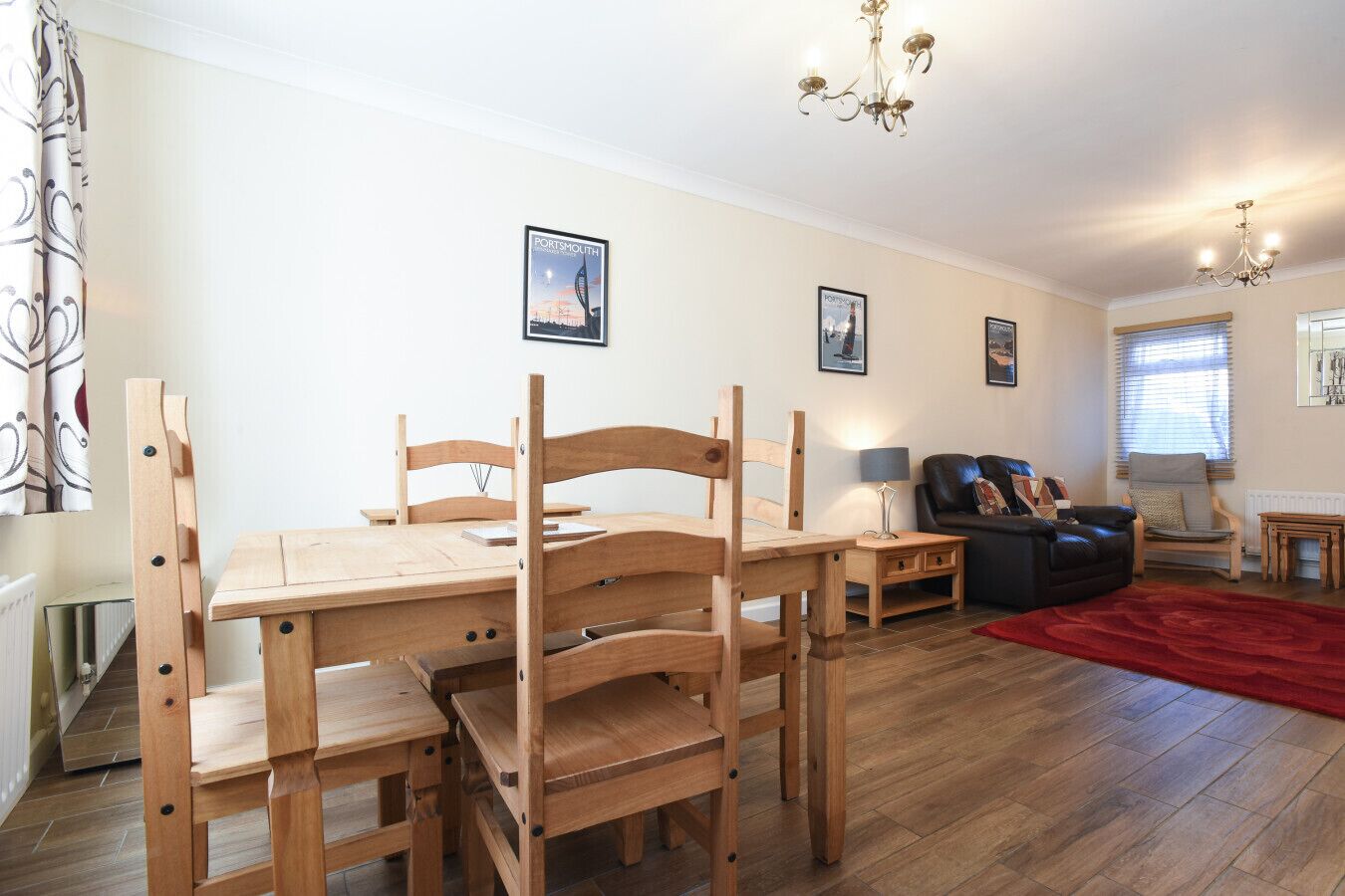 Ein gemütliches Wohnzimmer mit einem Holzesstisch und -stühlen, einer schwarzen Ledersofa und einem roten Teppich. An den Wänden hängen Bilder und von der Decke hängt eine Kristallleuchter.