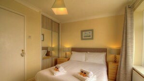 A hotel room with a large bed, bedside tables with lamps, a mirror, a window with curtains, and a painting on the wall. - Sea Breeze Haven | 4 Beds with Stunning Views! (Hayling Island)