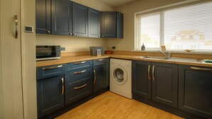 A compact kitchen with dark cabinets, a microwave, a washing machine, and a window with blinds. - Sea Breeze Haven | 4 Beds with Stunning Views! (Hayling Island)