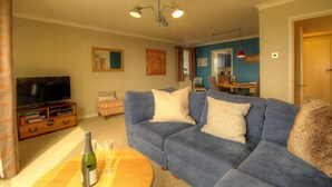 A living room with a blue sofa, a television on a wooden stand, a small table with a bottle and glasses, and a dining area in the background. - Sea Breeze Haven | 4 Beds with Stunning Views! (Hayling Island)