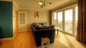 A living room with a dark-colored sofa, a small wooden stool, a table, and a painting on the wall. - Sea Breeze Haven | 4 Beds with Stunning Views! (Hayling Island)