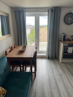 A dining area with a wooden table and chairs, a teal sofa, a kitchen area with a clock, and a view through a glass door.