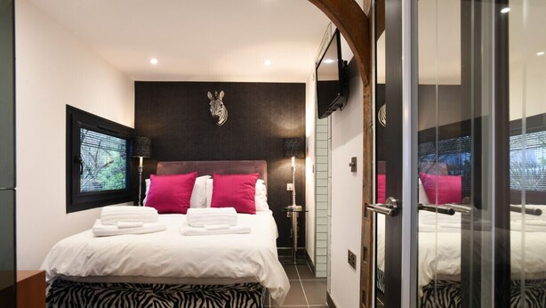 Ein modernes Hotelzimmer mit einem großen Bett, einem osmanischen Hocker mit Zebra-Muster, einer Zebra-Kopf-Dekoration an der Wand und einer Glastür, die in einen weiteren Raum führt.
