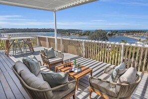 A wooden deck with wicker furniture, a table with drinks, and a view of a coastal town. - A Short Time - 2 Bedroom House (Merimbula)