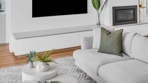 Un salon moderne avec un canapé blanc, une table basse ronde agrémentée de plantes décoratives, une cheminée et un grand téléviseur à écran plat fixé au mur.