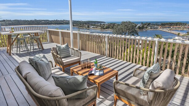 Une terrasse en bois avec des meubles en osier, une table avec des boissons et une vue sur une ville côtière.