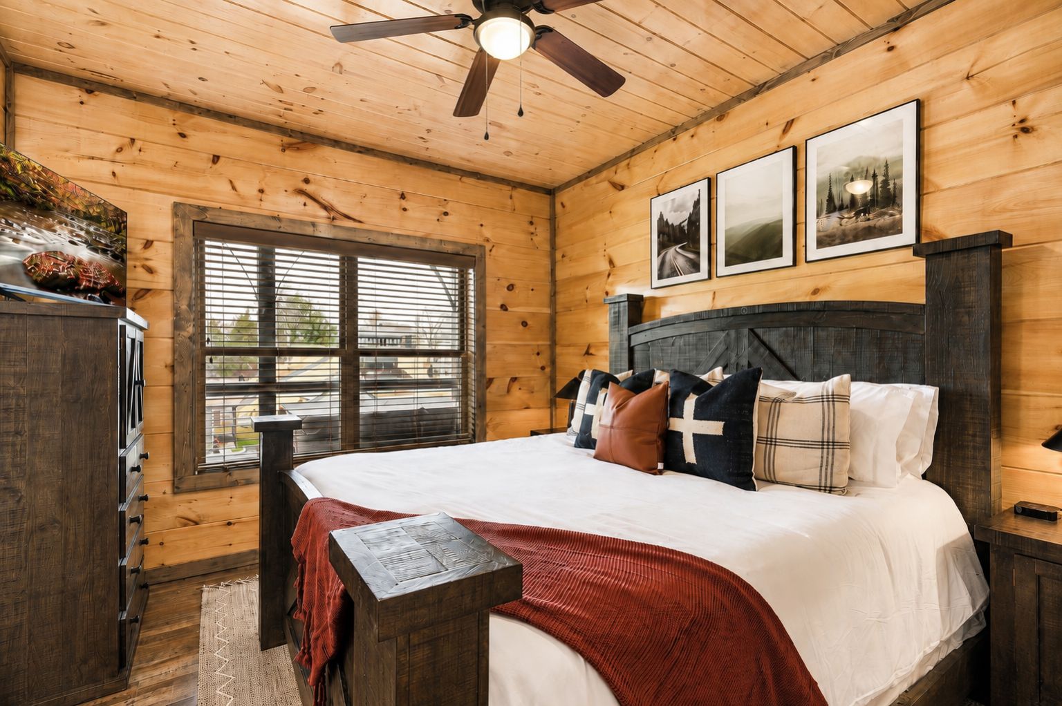 A wooden cabin bedroom with a bed, a ceiling fan, a window with blinds, and framed pictures on the wall.