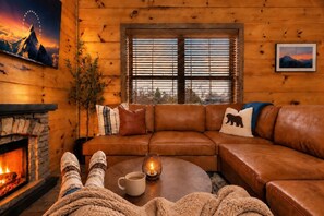 A cozy log cabin interior with a leather sofa, a fireplace, and a window with blinds.
