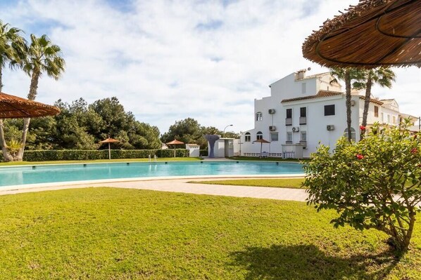 A pool area with a white building, palm trees, and a thatched umbrella.