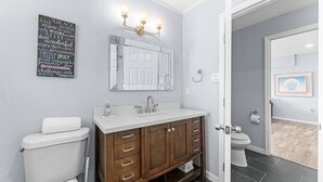 A bathroom with a white toilet, a wooden vanity with a mirror, and a wall-mounted light fixture. - 🐾  Dog Friendly, Spacious Beach Retreat, only Steps to Sugary Sand, Conveniences and all the Fun! (Fort Myers Beach)