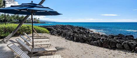 A beachside lounge with wooden lounge chairs and a blue umbrella, overlooking a rocky shoreline and the ocean.