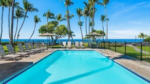 A pool area with lounge chairs and umbrellas, surrounded by palm trees and a view of the ocean. - 🏖️Ocean Front! | Second Floor with AC & Lanai! (Kailua Kona)