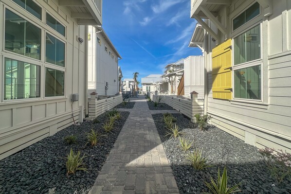 A narrow pathway flanked by white buildings with large windows and a yellow door, leading to a central area with a paved walkway and small plants. - Resort Pools, Golf Cart Boardwalk to Beach (Port Aransas)