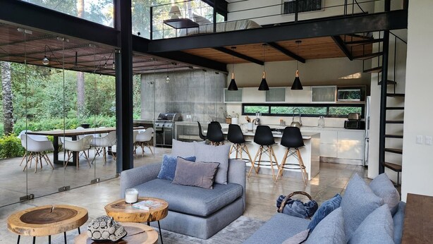 Un espacio habitable moderno y abierto con un sofá gris, mesas de madera redondas y taburetes negros.