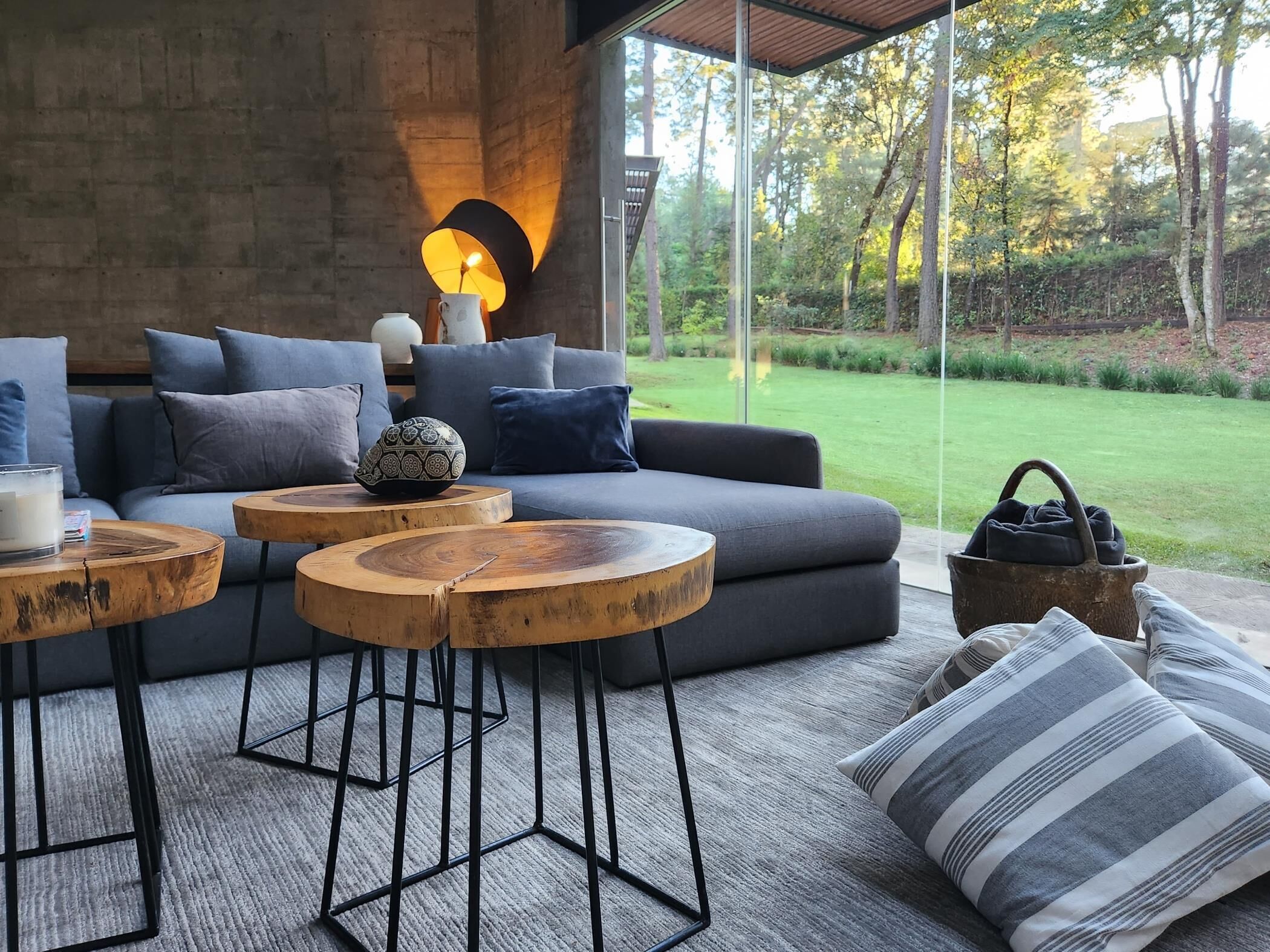 Een moderne woonkamer met een grijze bank, ronde houten tafels en een groot raam met uitzicht op een tuin.