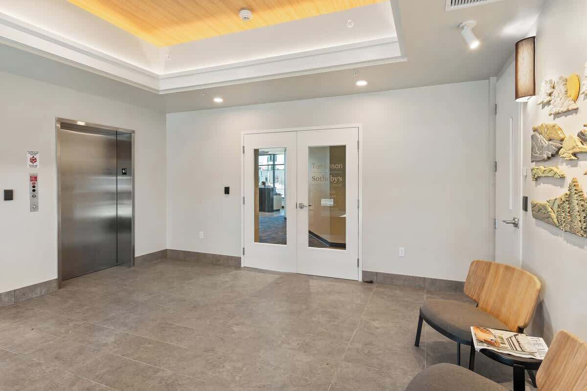 A modern lobby with a reception desk, a waiting area with chairs, a glass door entrance, and a staircase leading to an elevator.