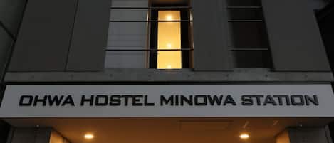 The exterior of Ohwa Hostel Minowa Station, featuring a modern architectural design with a prominent entrance and illuminated signage.