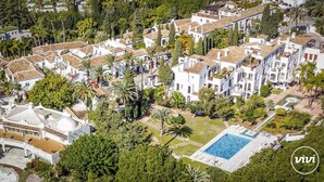 Oversigtsbillede af et boligområde med hvide bygninger, en swimmingpool og frodig beplantning.