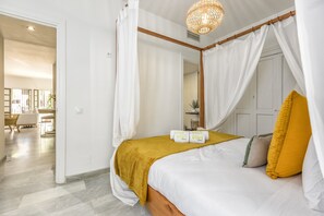 A four-poster bed with a white canopy, a mustard-colored throw blanket, and two rolled towels with a logo.