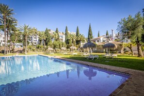 A swimming pool with lounge chairs and umbrellas, surrounded by greenery and buildings.