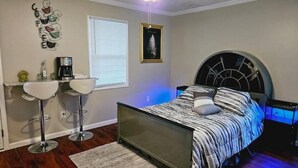 A bedroom with a bed, a coffee maker, two chairs, a window, and a framed picture on the wall. - Studio Kevdaya 1 (Norcross)