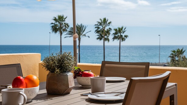 A table with a laptop, a bowl of fruit, a potted plant, and a white mug, set against a backdrop of the ocean and palm trees.