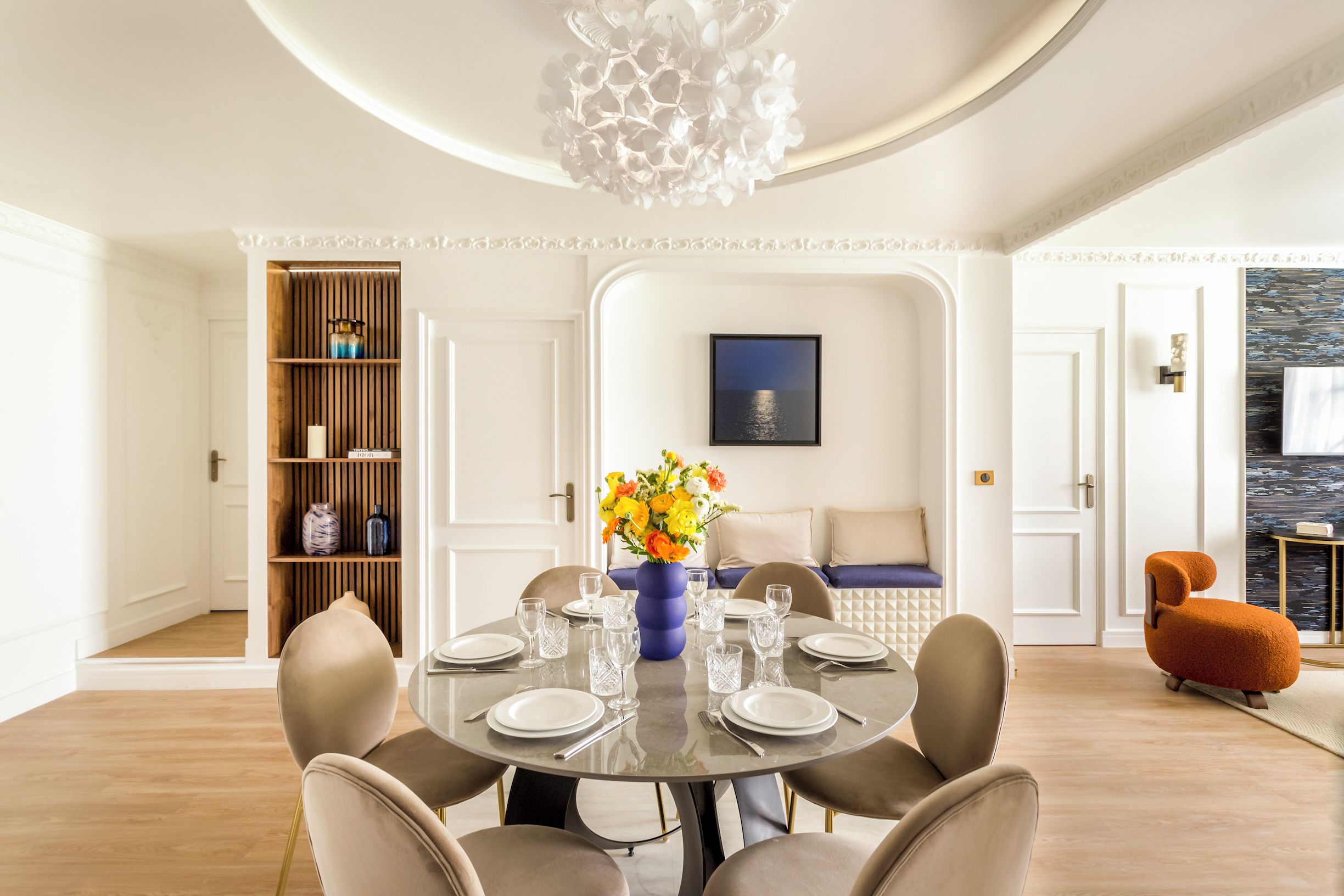 A dining area with a round table set for six, surrounded by six chairs. There is a blue vase with flowers on the table, and a wooden bookshelf to the left.