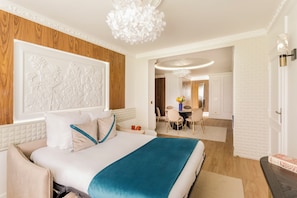 A modern hotel room with a large bed, a wooden accent wall, a chandelier, and a dining area visible through an open door.