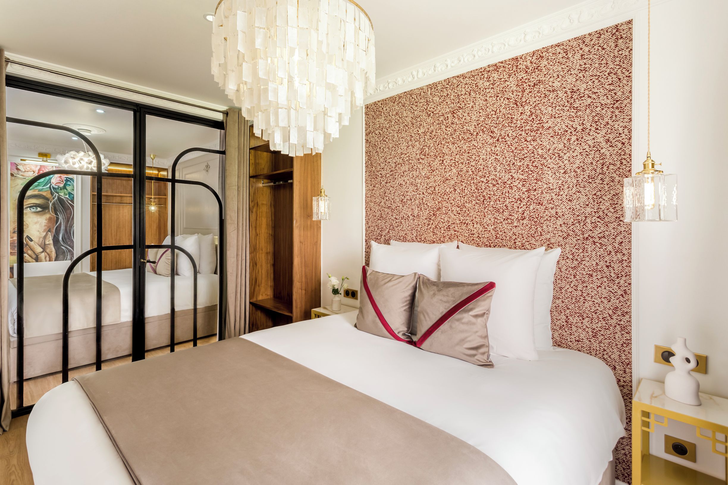 A hotel room with a large bed, a chandelier, a floral-patterned wall, and a sliding door leading to a bathroom.