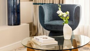 Una sala de estar moderna con una mesa de centro de vidrio, un sillón azul oscuro, un jarrón blanco con flores y un jarrón azul alto frente a una pared beige.