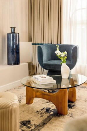 A modern living room with a glass coffee table, a dark blue armchair, a white vase with flowers, and a tall blue vase against a beige wall. - Merveil - Luxury Suite - Marais - Lenoir III (Paris)