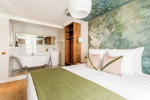 A hotel room with a large bed, a green throw blanket, a wooden wardrobe, a marble bathroom, and a wall with a floral mural.