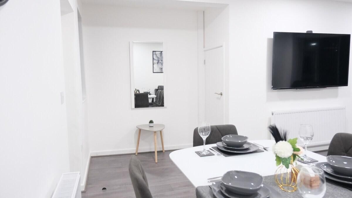 A modern dining area with a white table set for four, a small side table, and a black TV mounted on the wall.