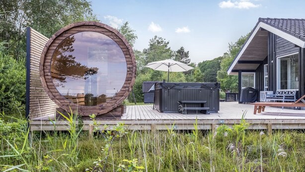 Una terraza de madera con un jacuzzi, mobiliario exterior y un gran ventanal circular que refleja el cielo.