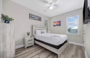 A bedroom with a bed, a nightstand, a ceiling fan, a window with blinds, and a wall-mounted TV. - Coastal Home 3BR-6 Beds EV Charger+ Garage•Walk to Beach (Myrtle Beach)
