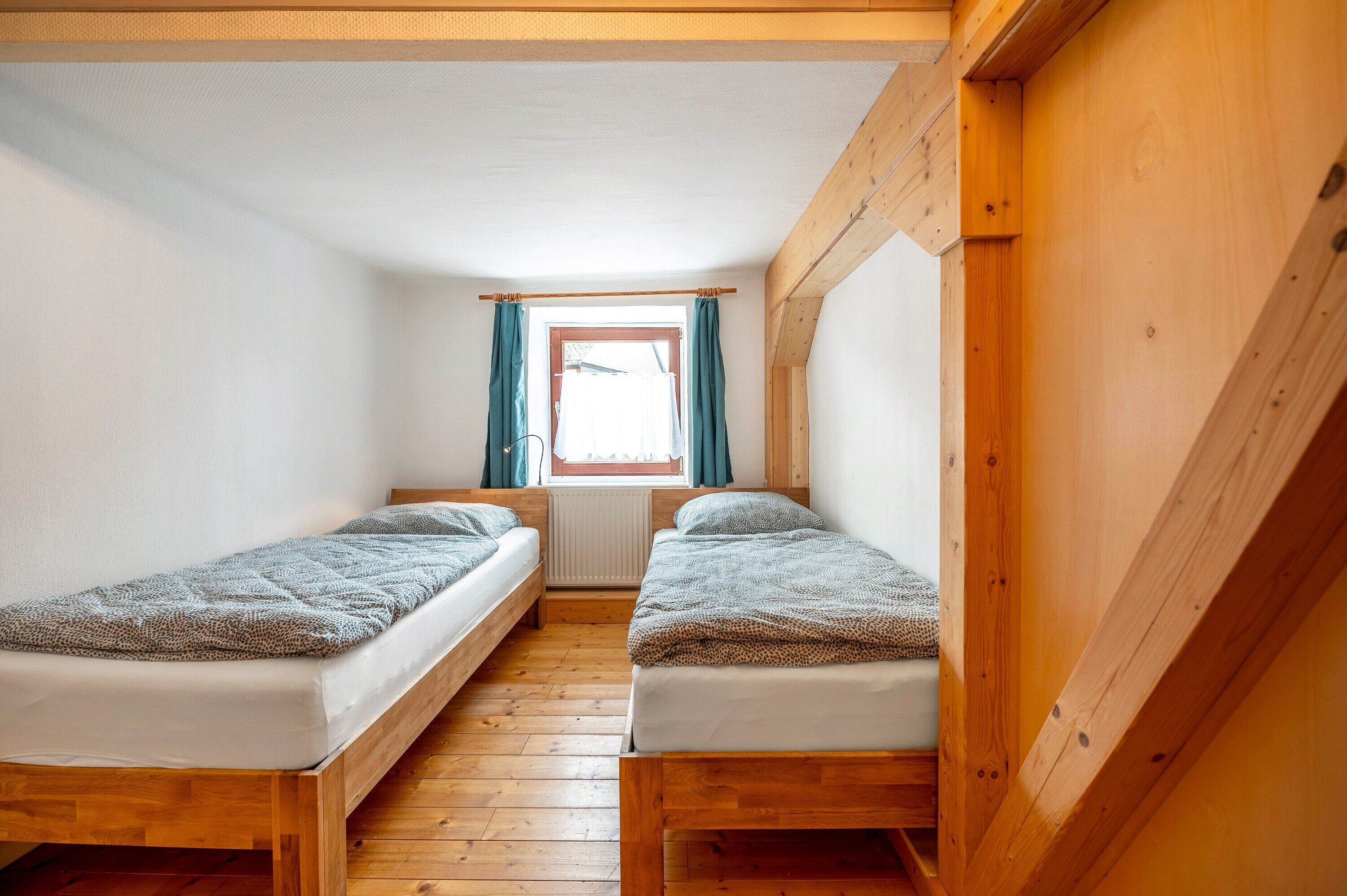 Een kleine kamer met houten wanden, twee enkele bedden, een raam met gordijnen en een radiator.