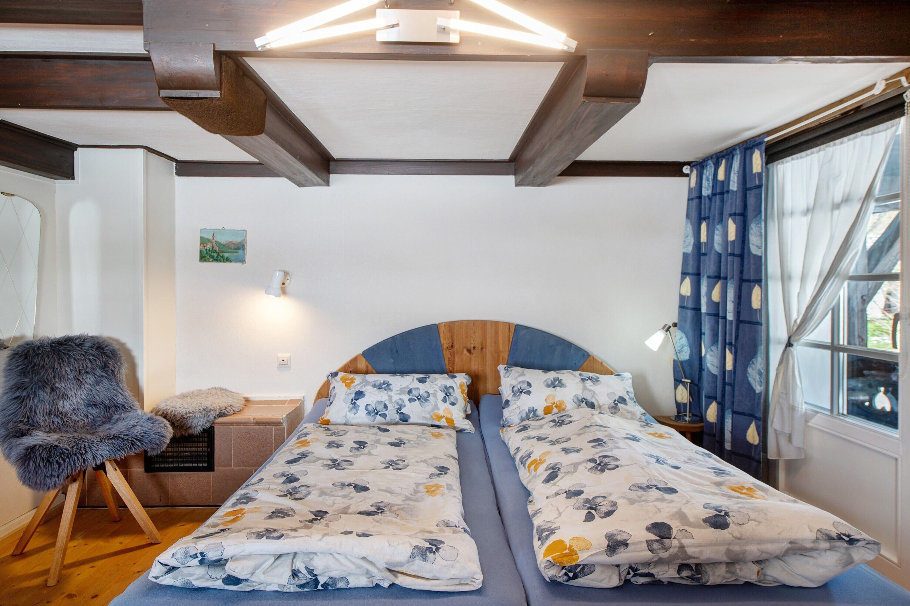 A bedroom with a wooden headboard, two single beds with floral bedding, a chair with a fur throw, a bedside table with a lamp, and a window with curtains.