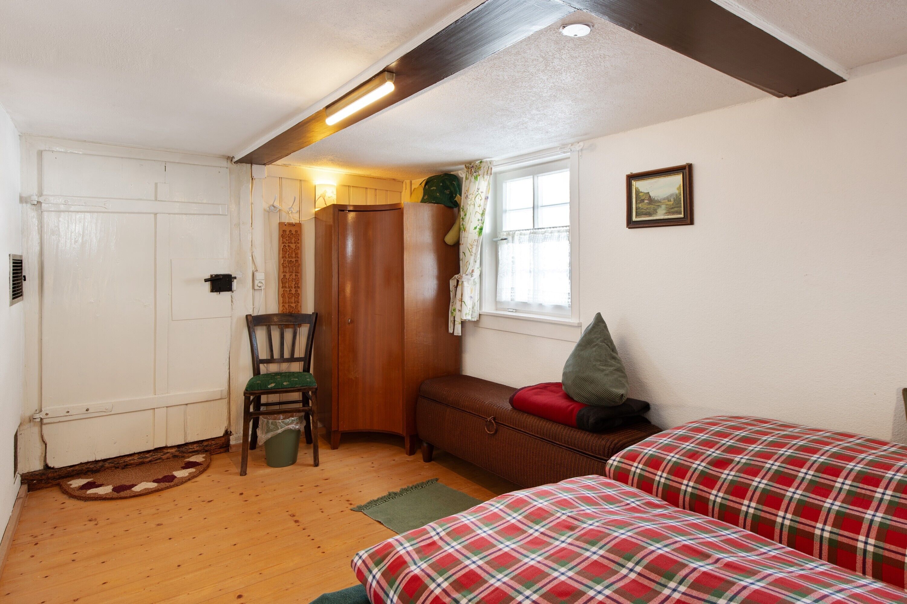 A room with a wooden floor, a bench with a red cushion, a chair with a green cushion, a wooden wardrobe, a small window with a curtain, and a framed picture on the wall.