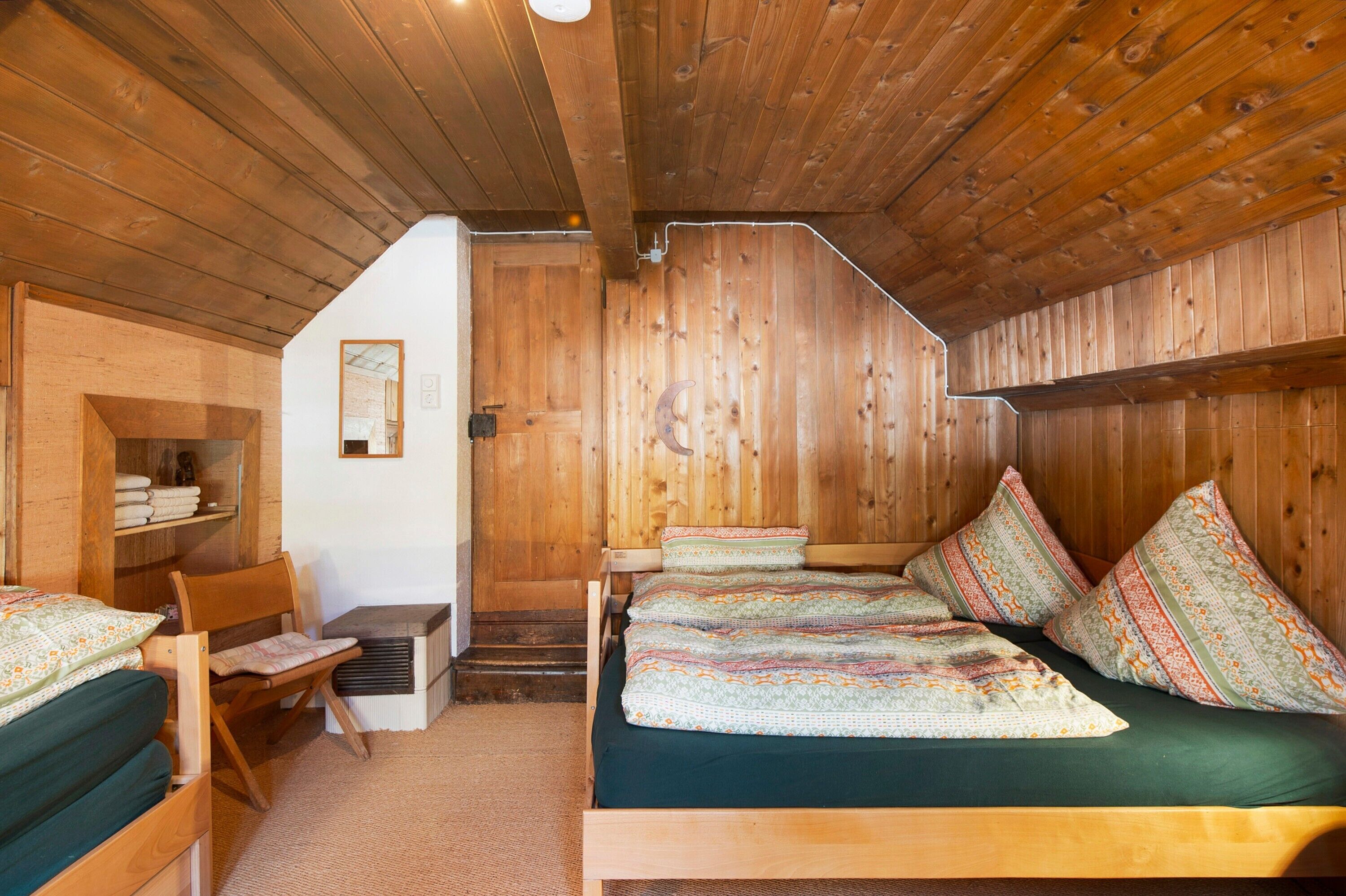 A wooden cabin interior with a loft bed, a chair, a small table, and a staircase leading to an upper level.