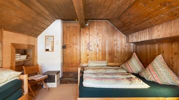 A wooden cabin interior with a loft bed, a chair, a small table, and a staircase leading to an upper level.