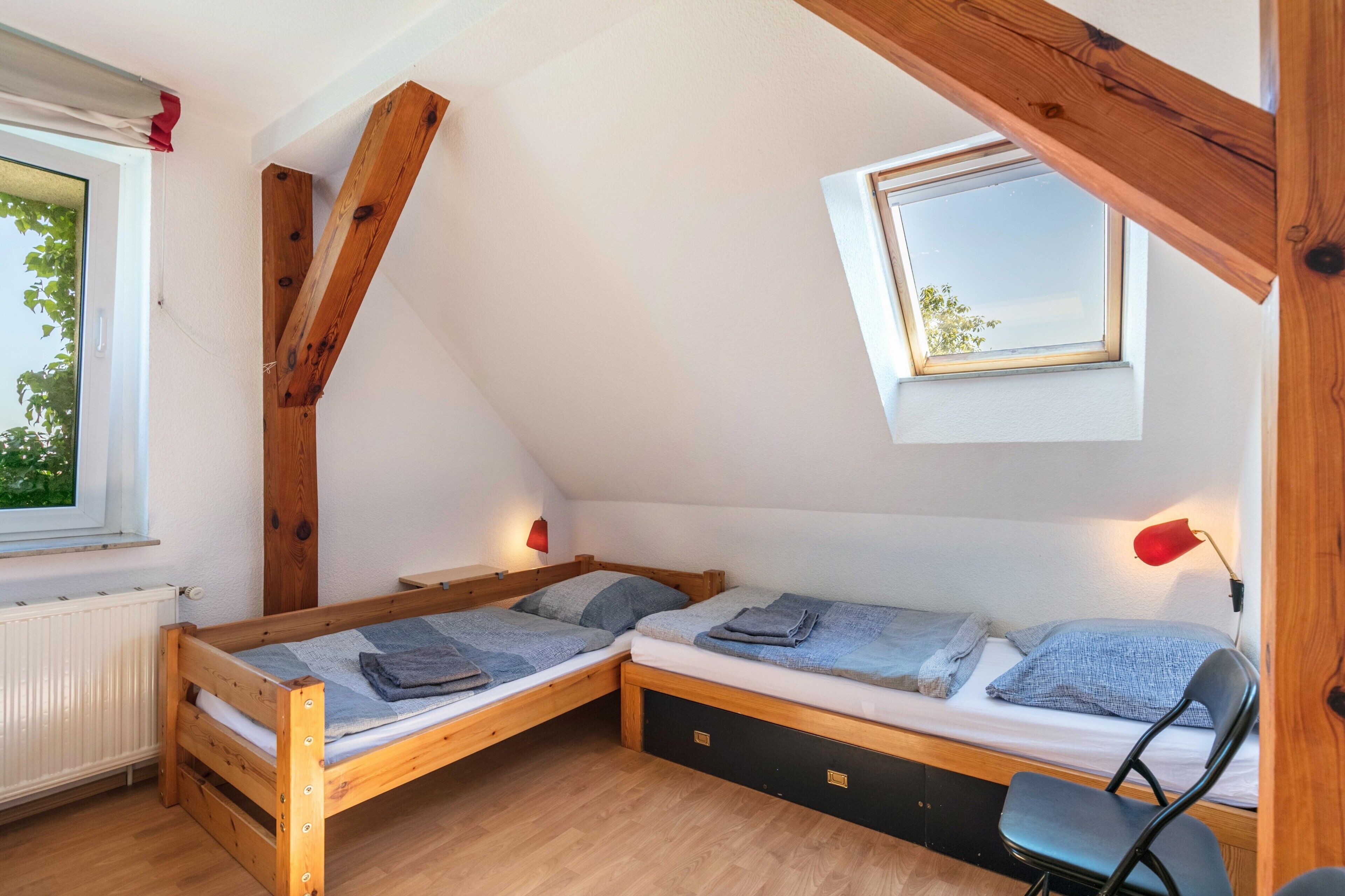 Una habitación pequeña y acogedora con dos camas de madera, una ventana, un tragaluz y una silla.