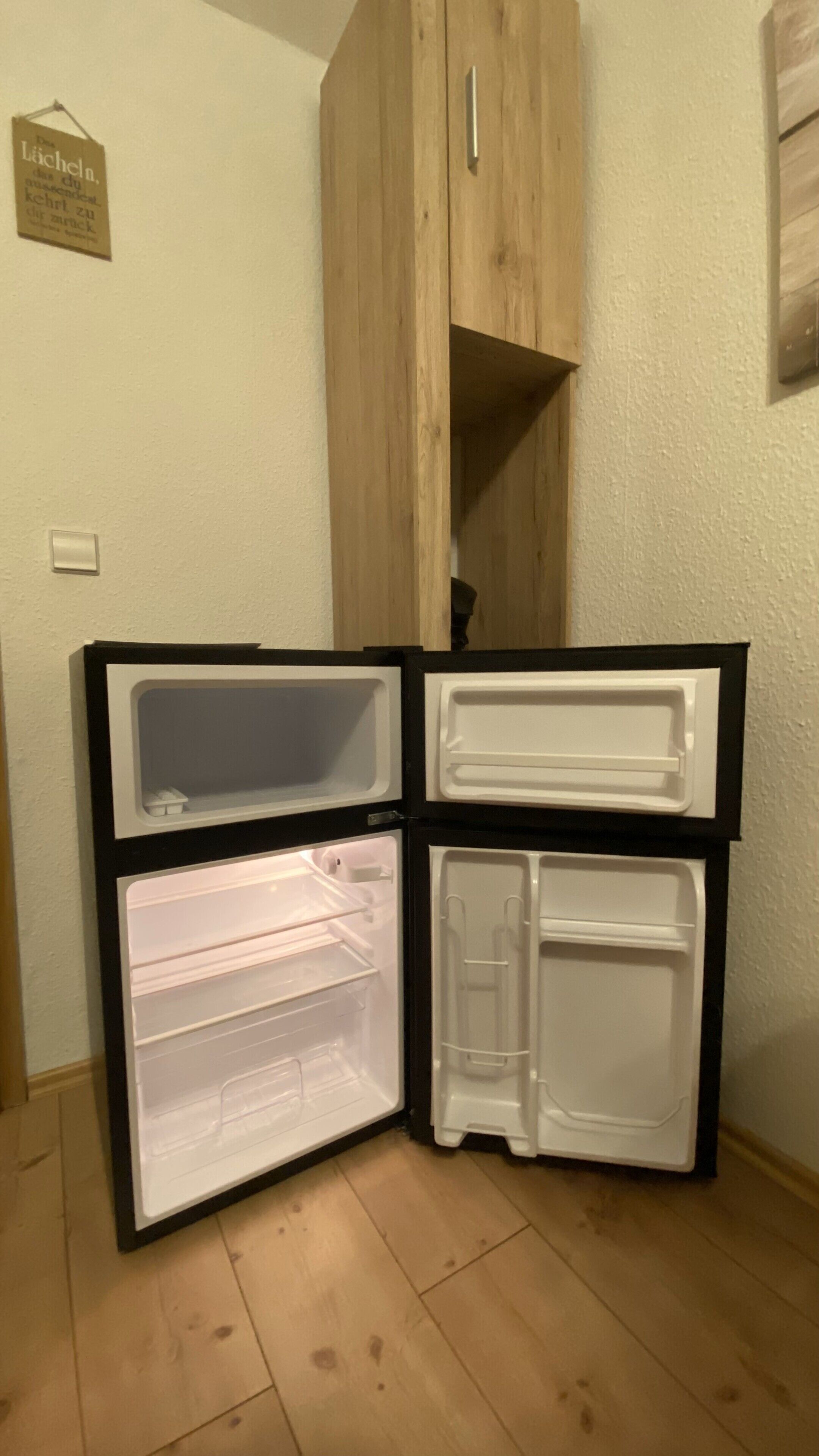 Ein kompakter Kühlschrank mit zwei Fächern in der Tür und einer Gefrierablage, platziert in einer Ecke mit Holzboden und weißer Wand.