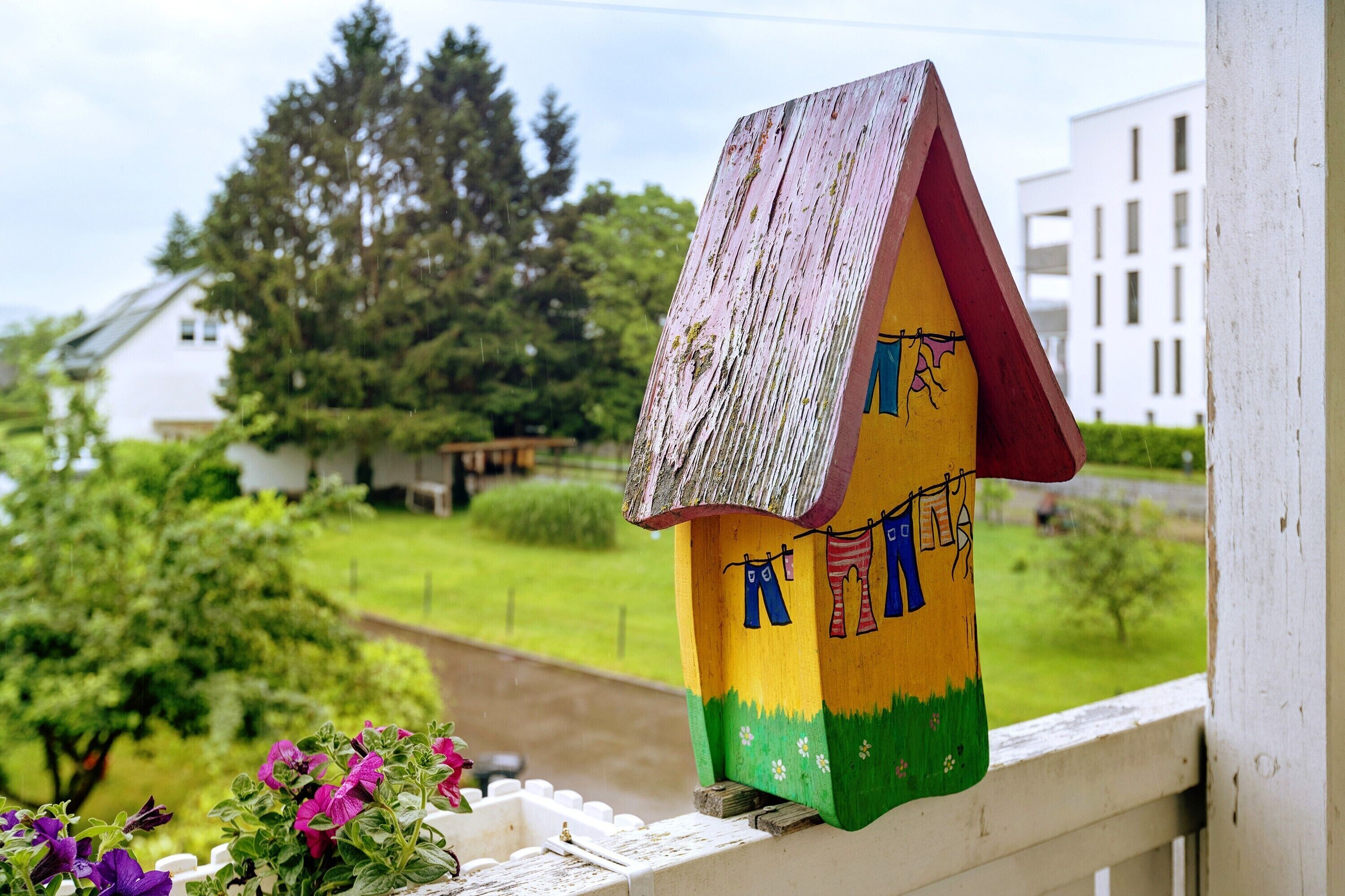 Una casa para pájaros con techo desgastado y decoraciones coloridas, ubicada en un balcón con vistas a un paisaje verde y edificios.