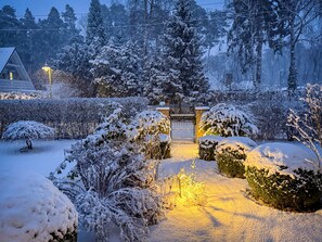 Un jardin enneigé, avec un chemin illuminé menant à une porte, entouré d’arbres chargés de neige et d’un bâtiment visible au loin.