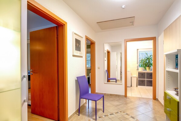 Un pasillo con una puerta de madera, una silla morada, un espejo y una planta junto a una ventana.