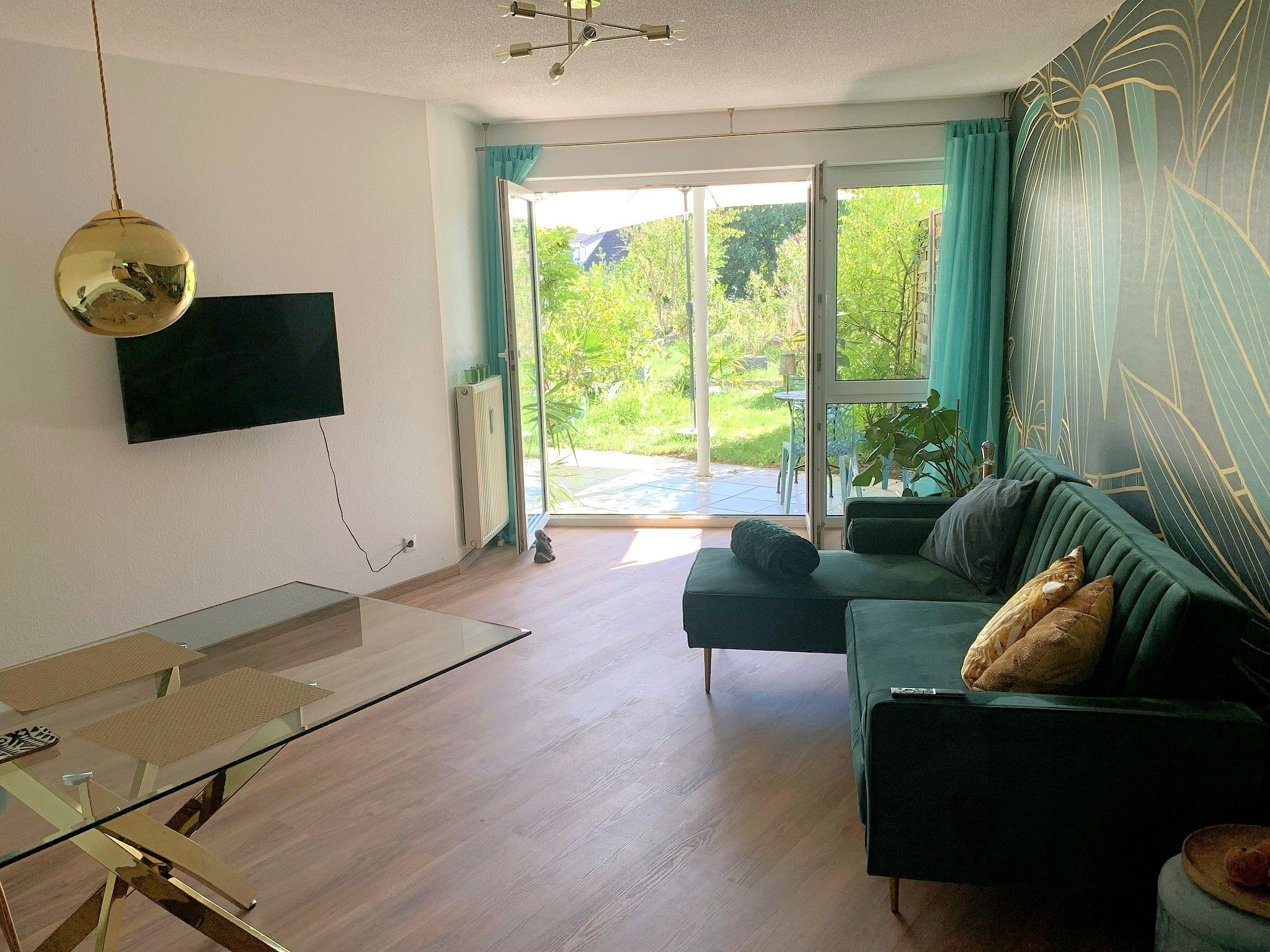 Un salon avec un canapé vert, un téléviseur à écran plat fixé au mur et une table basse en verre.