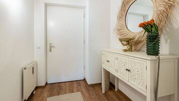 Um corredor com uma porta branca, um radiador, um tapete estampado, um console branco com detalhes entalhados e um espelho com moldura decorativa.