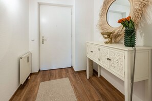 Een hal met een witte deur, een radiator, een kamerbreed tapijt met een patroon, een witte console met ingewikkelde gravures, en een spiegel met een decoratieve lijst.