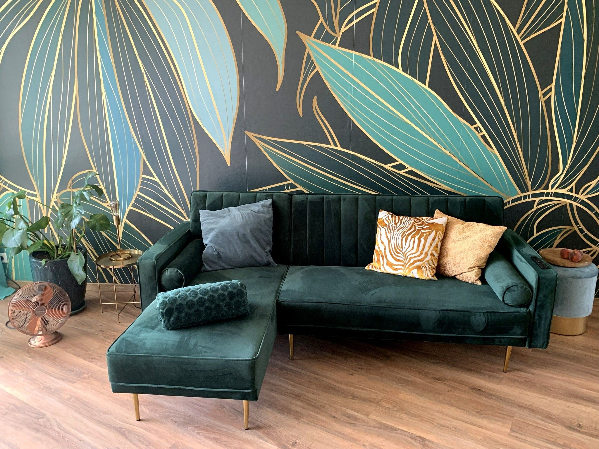 Ein modernes Wohnzimmer mit einem dunkelgrünen Ecksofa, einem kleinen Hocker und einer großen Wandmalerei mit abstrakten Blattmustern.