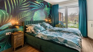 Makuuhuoneessa on suuri sänky, yöpöytä, seinämaalaus ja ikkuna, jossa on verhot.